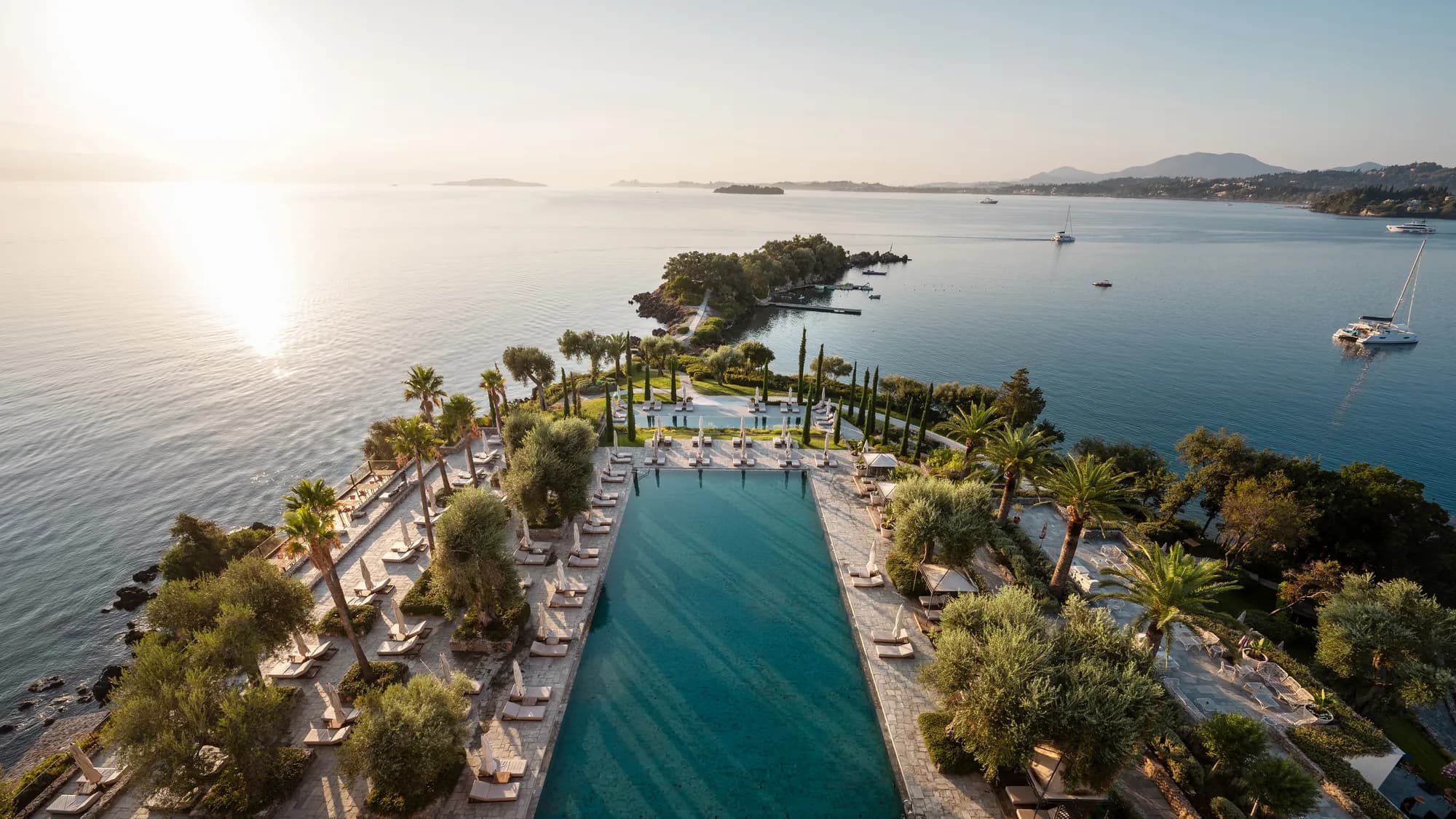 Das lange Becken des Corfu Imperial zwischen Palmen im Abendlicht, dahinter das Meer